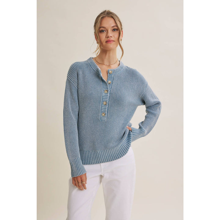 A woman modeling a light blue ribbed knit sweater with a buttoned henley neckline, paired with white pants, against a plain beige studio background.