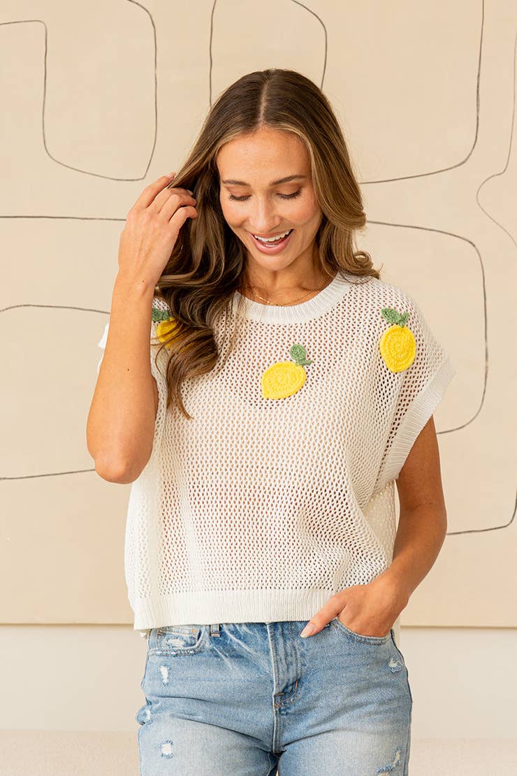 Woman wearing a sleeveless white top with lemon patterns against a beige wall.