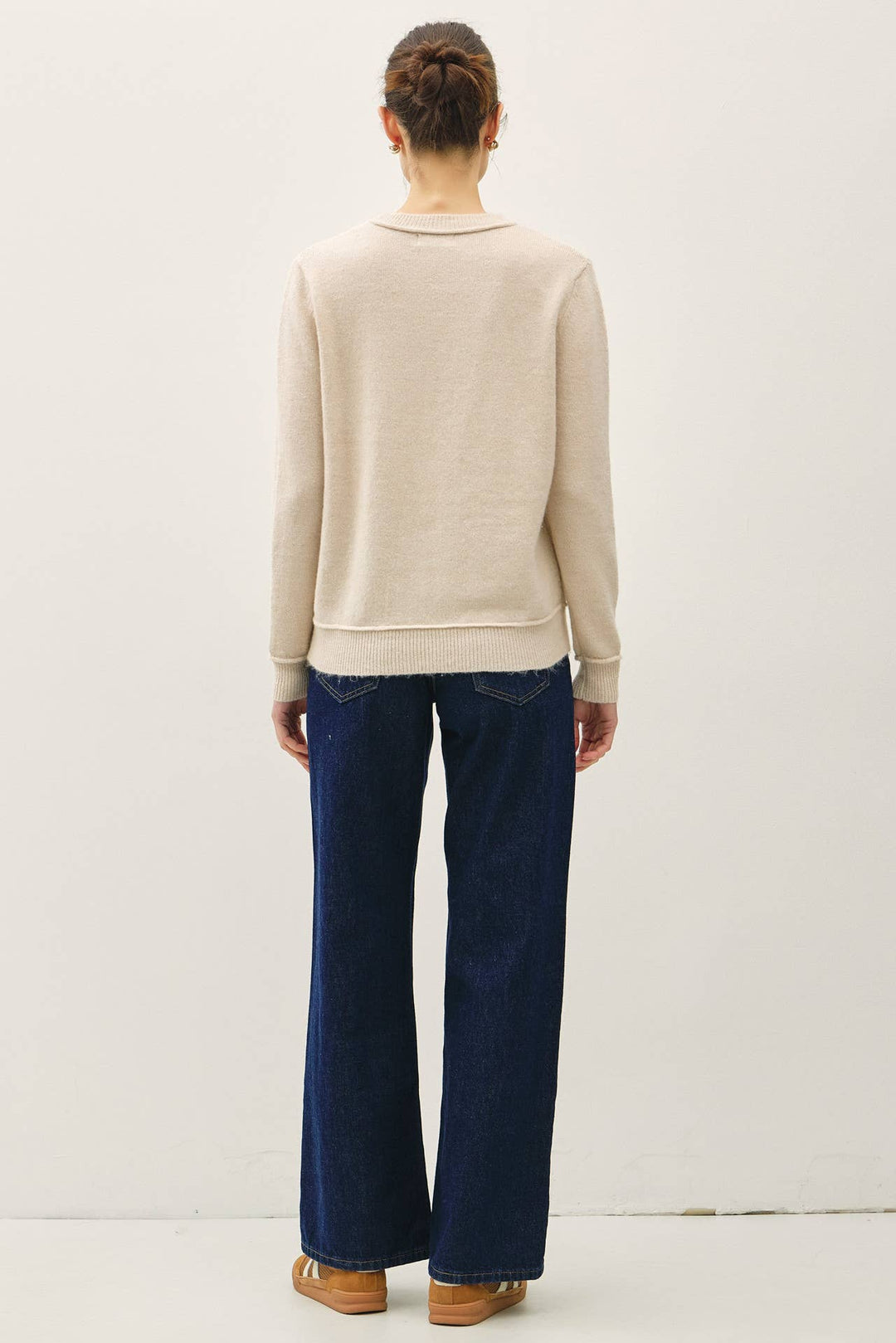 A model shown from the back wearing a light beige crewneck sweater and dark blue wide-leg jeans, standing against a plain off-white wall.