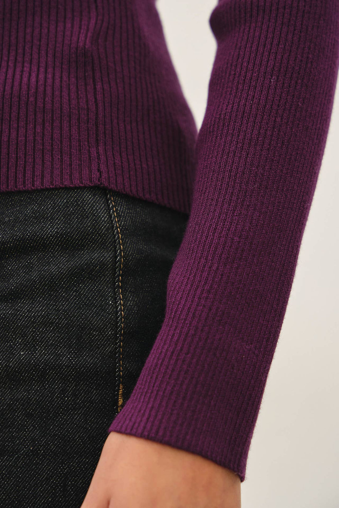Close-up of a person wearing a ribbed long-sleeve knit top in deep purple with dark denim jeans, showing the sleeve cuff and part of the torso against a plain light background.
