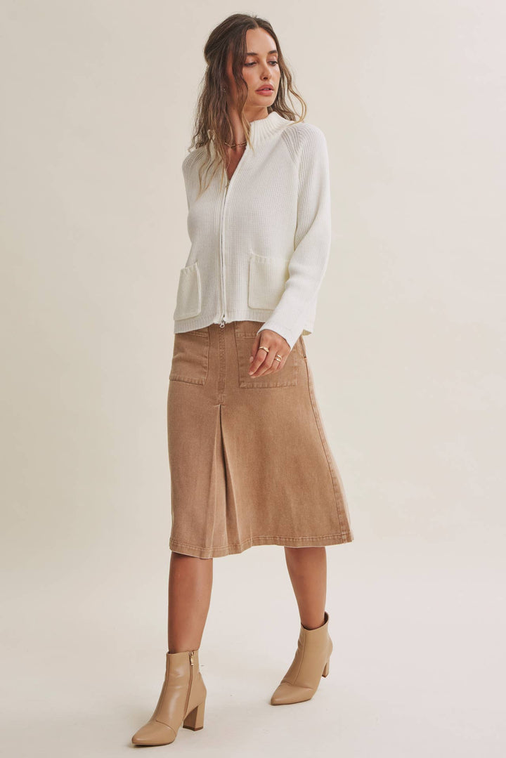 A woman modeling a white knit zip-front cardigan with front patch pockets, paired with a tan corduroy midi skirt and beige ankle boots, against a plain light beige studio background.