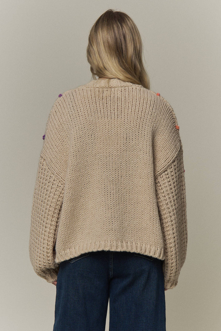 A woman viewed from behind wearing a chunky knit beige sweater with colorful  details on the shoulders and dark blue jeans, set against a plain light gray studio background.