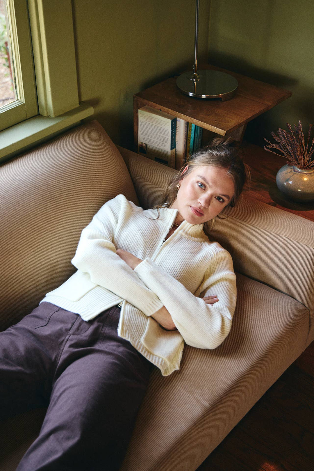 Woman sitting on a couch in a cozy room with a lamp and window in the background.