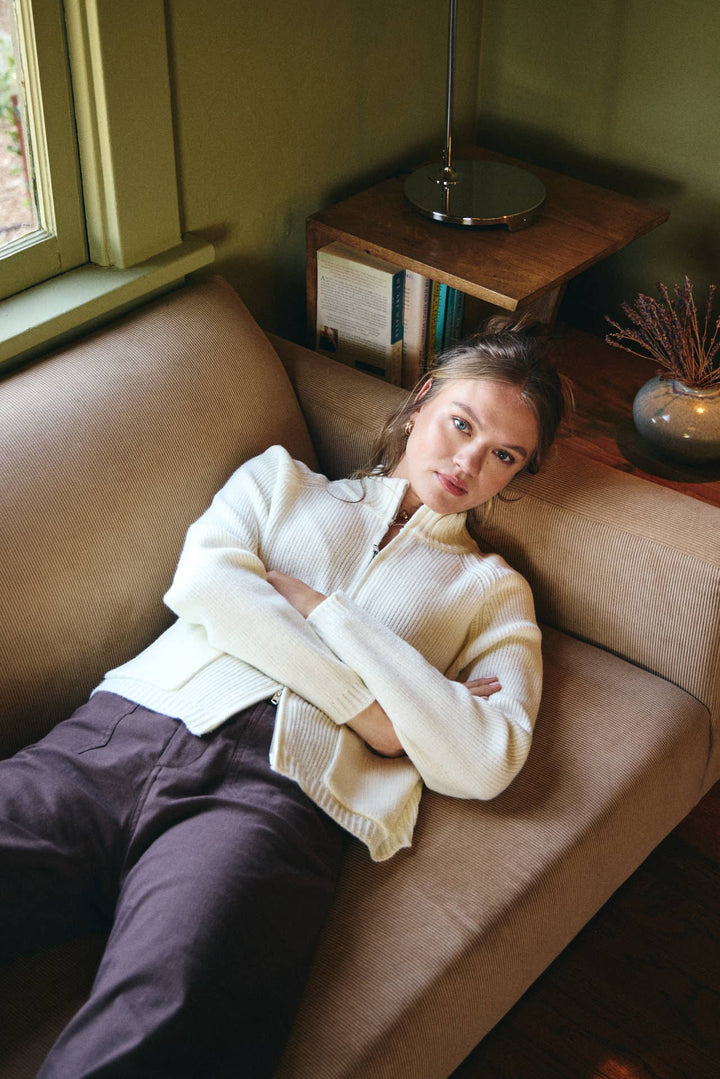 Woman sitting on a couch in a cozy room with a lamp and window in the background.