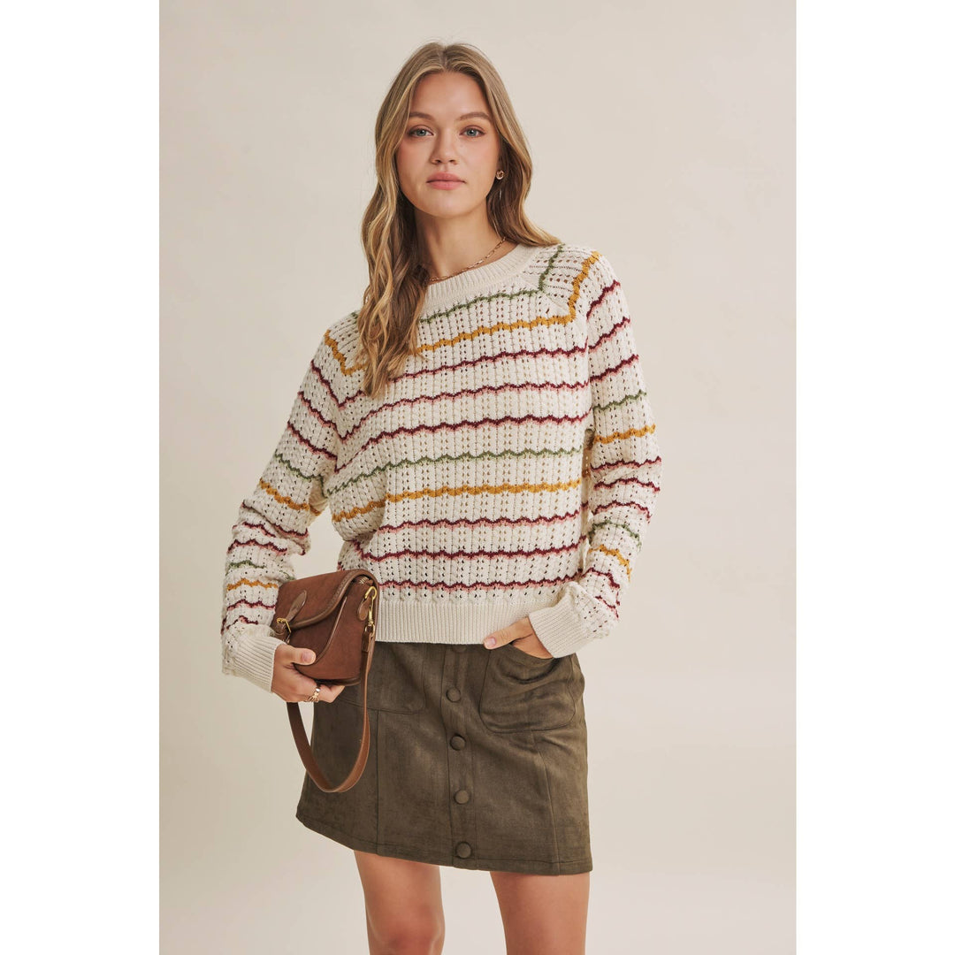 A woman modeling a cream knit sweater with horizontal stripes in red, yellow, and green, paired with an olive green mini skirt and holding a brown leather crossbody bag against a solid beige studio background.
