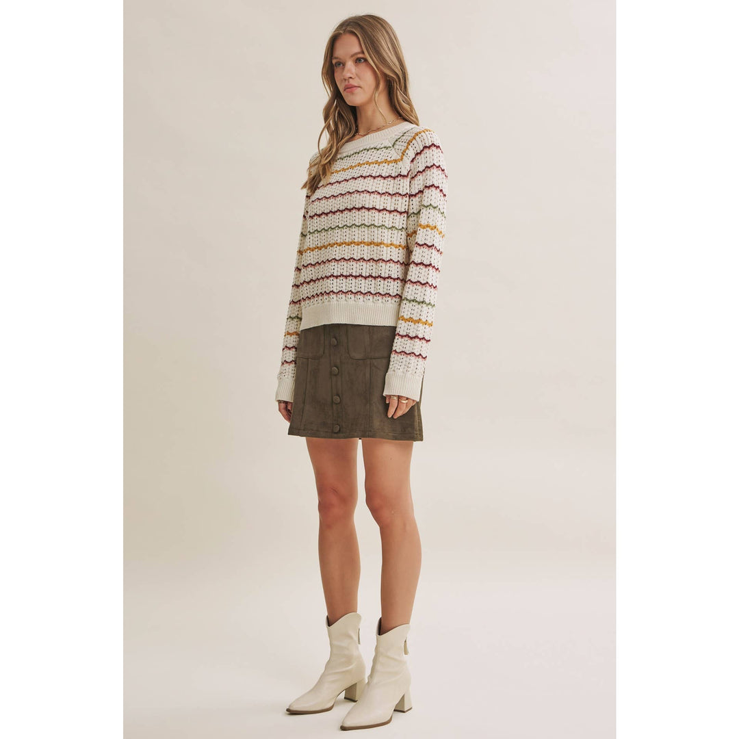 A woman modeling a cream knit sweater with horizontal multicolor stripes in red, yellow, and orange, paired with a dark brown mini skirt and off-white ankle boots, against a solid light beige studio background.