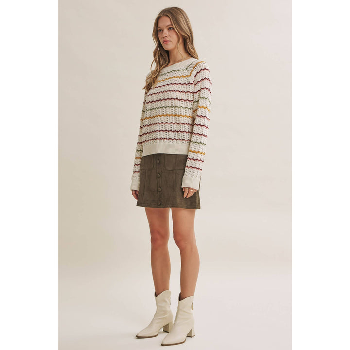A woman modeling a cream knit sweater with horizontal multicolor stripes in red, yellow, and orange, paired with a dark brown mini skirt and off-white ankle boots, against a solid light beige studio background.