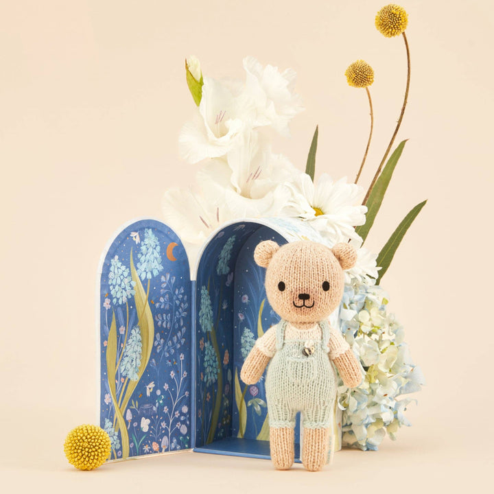 Knitted teddy bear in front of a floral arrangement and product packaging on a beige background