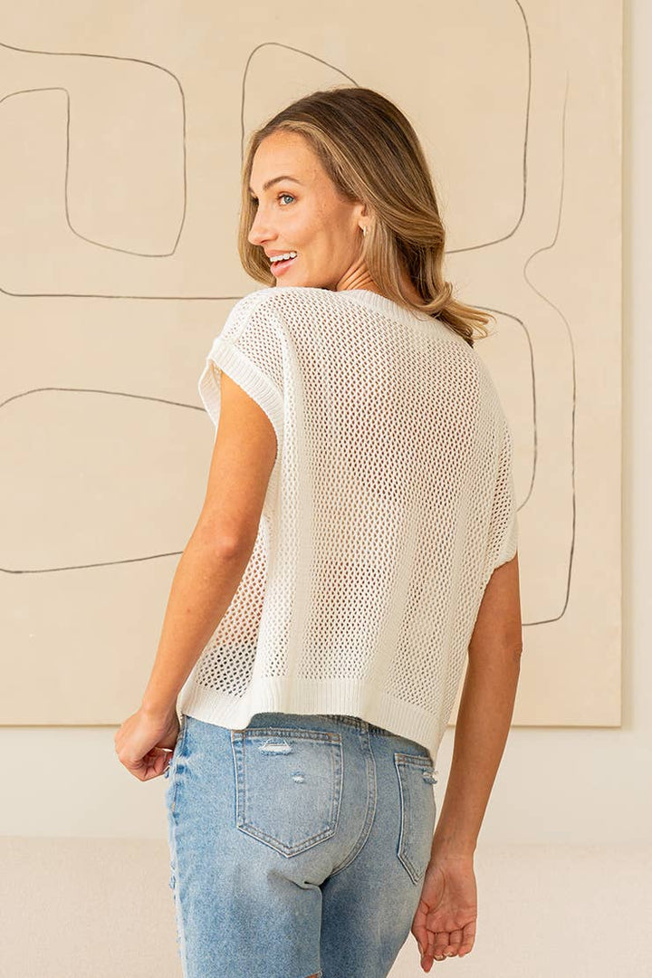 Woman wearing a beige knit top and blue jeans against a neutral wall.