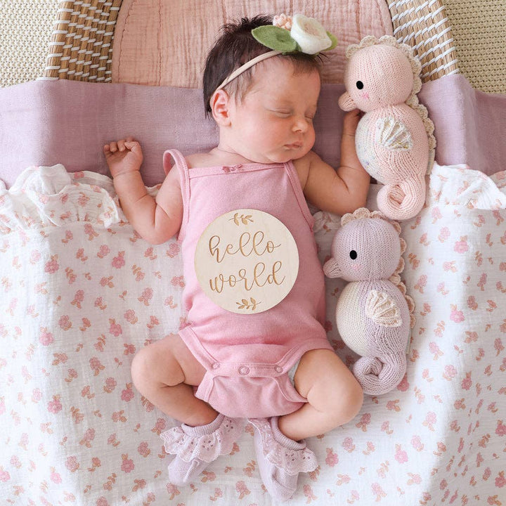 Baby in a pink 'hello world' outfit lying on a floral blanket with stuffed sea creature toys.