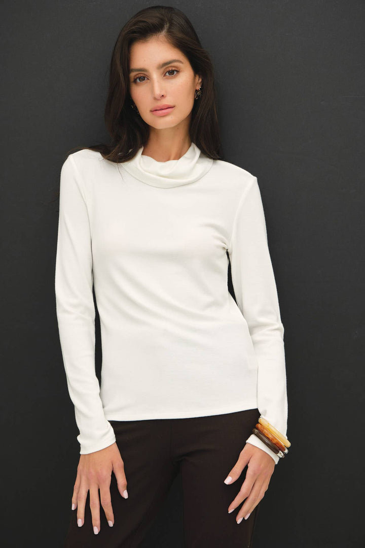 A woman wearing a white long-sleeve turtle neck top and dark brown pants, standing against a dark gray background.