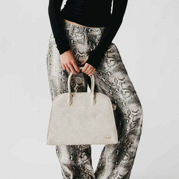 Person holding a oat colored handbag wearing patterned pants on a light gray background