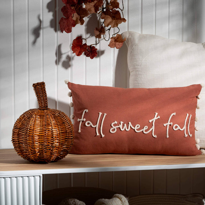 Decorative pillow with 'fall sweet fall' text on a wooden surface next to a wicker pumpkin.