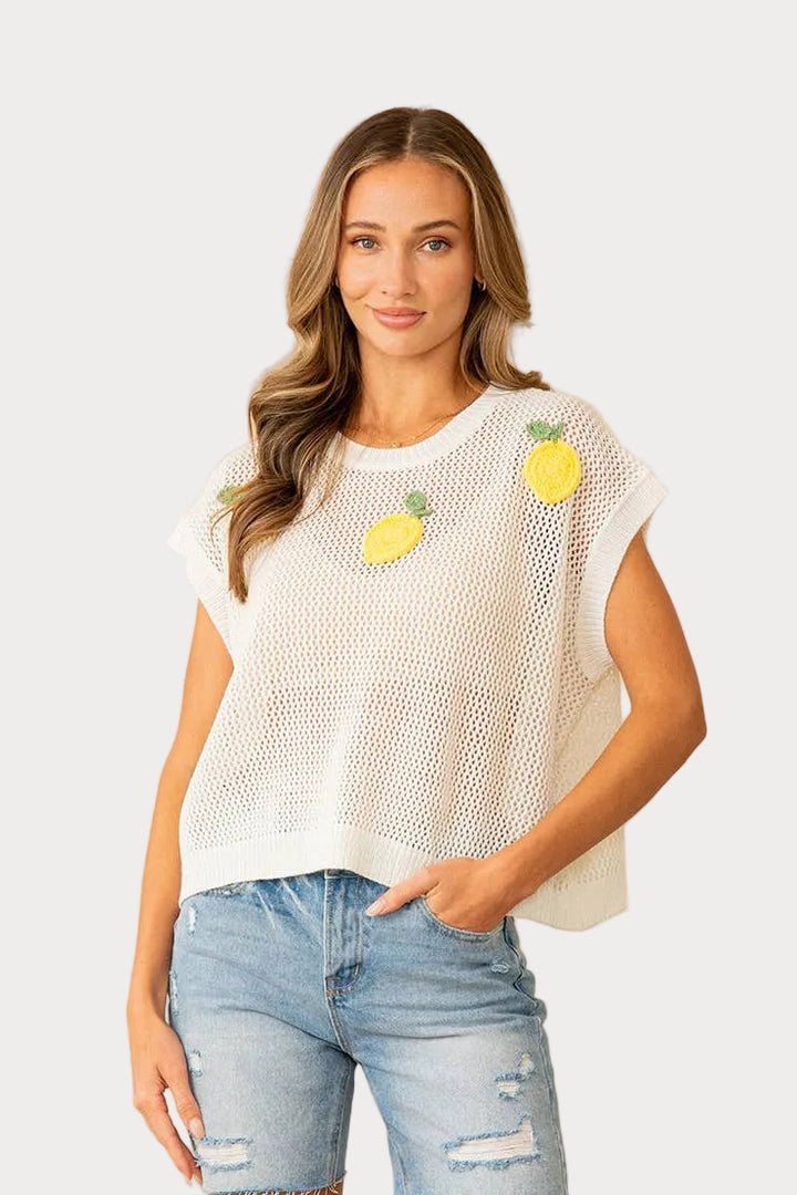 Woman wearing a white mesh top with lemon designs and light blue jeans against a beige wall.