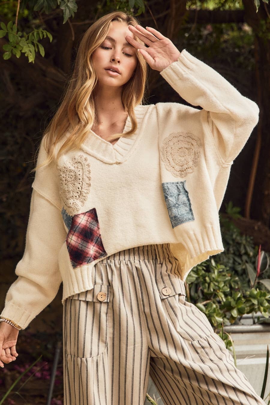 A woman wearing a cream knit sweater with crochet and patchwork details, paired with cream pinstriped trousers, posing outdoors in front of a garden background.
