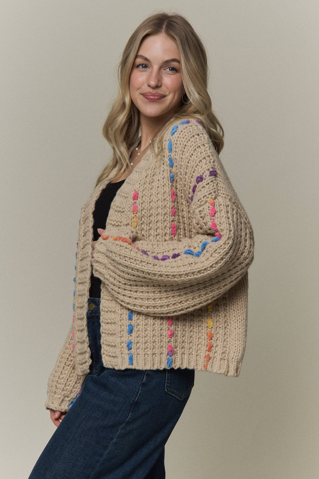 A woman wearing a chunky knit cardigan in a warm beige color with vertical and horizontal multicolor striped patterns in pink, blue, yellow, and purple. She is posing against a solid light beige background.