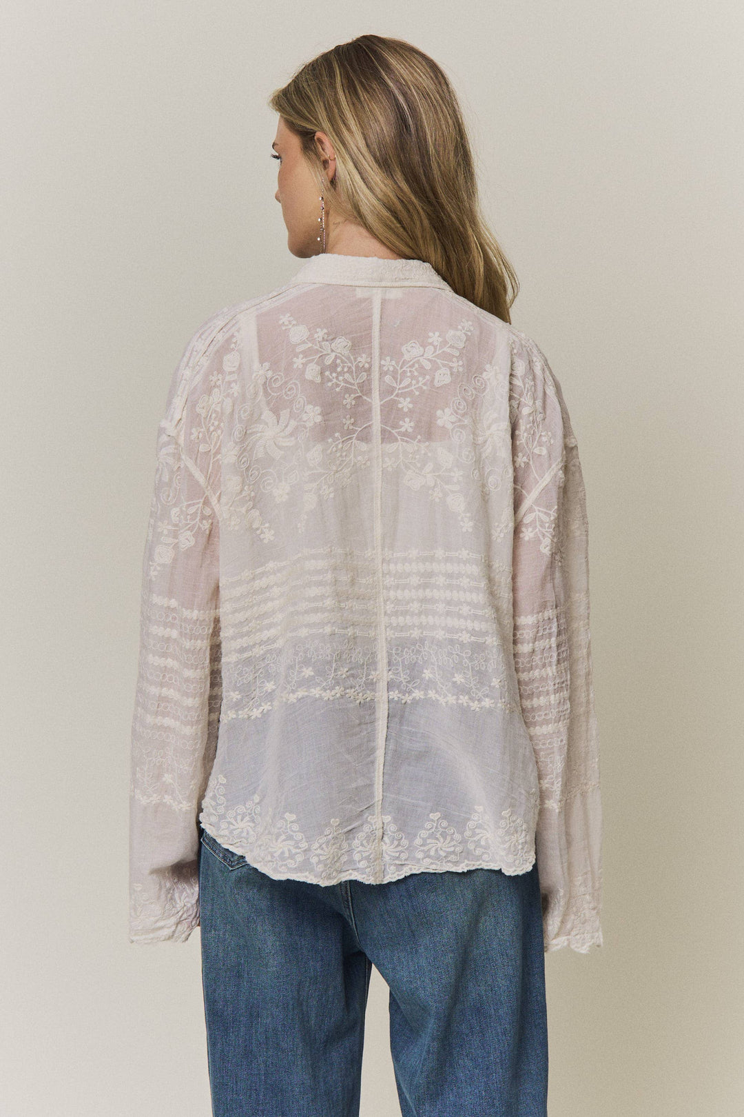 Woman wearing a light-colored lace embroidered blouse and blue jeans against a plain background