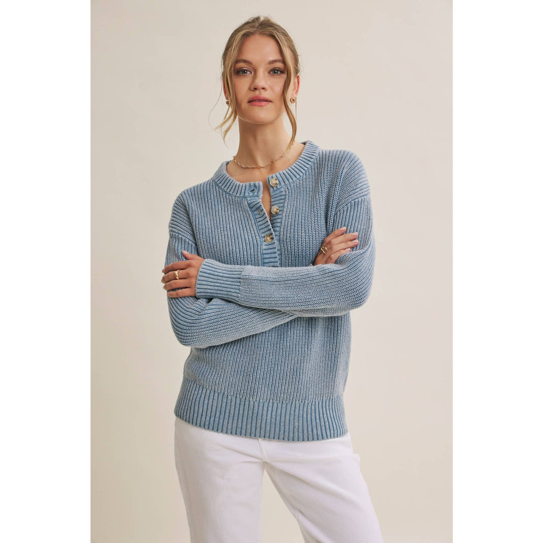 A woman modeling a light blue ribbed knit cardigan sweater with a buttoned henley neckline, paired with white pants, against a plain beige studio background.
