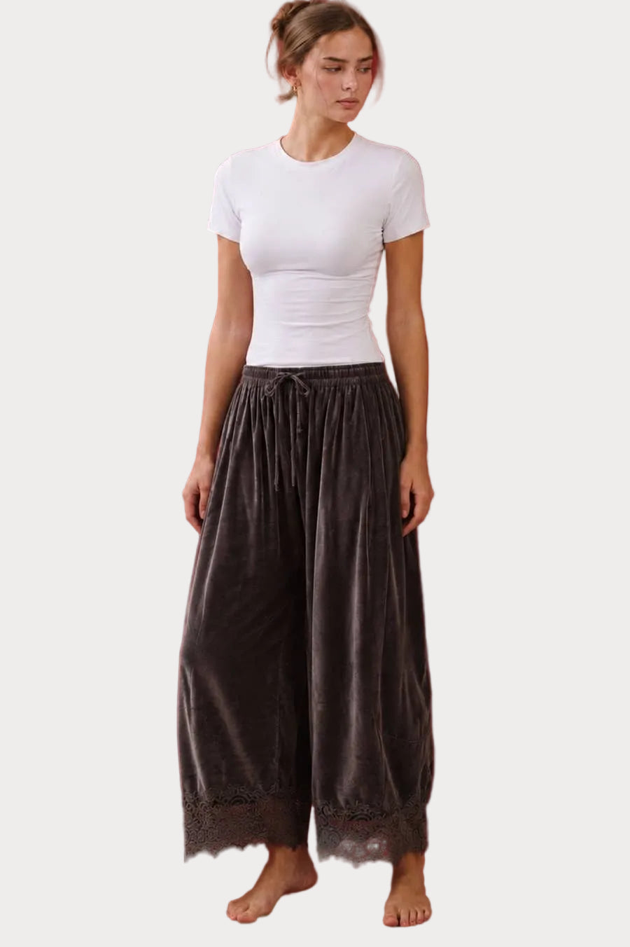 A woman stands barefoot against a solid studio background, wearing a fitted white short-sleeve top and loose dark brown gray velvet wide-leg pants with a lace hem.