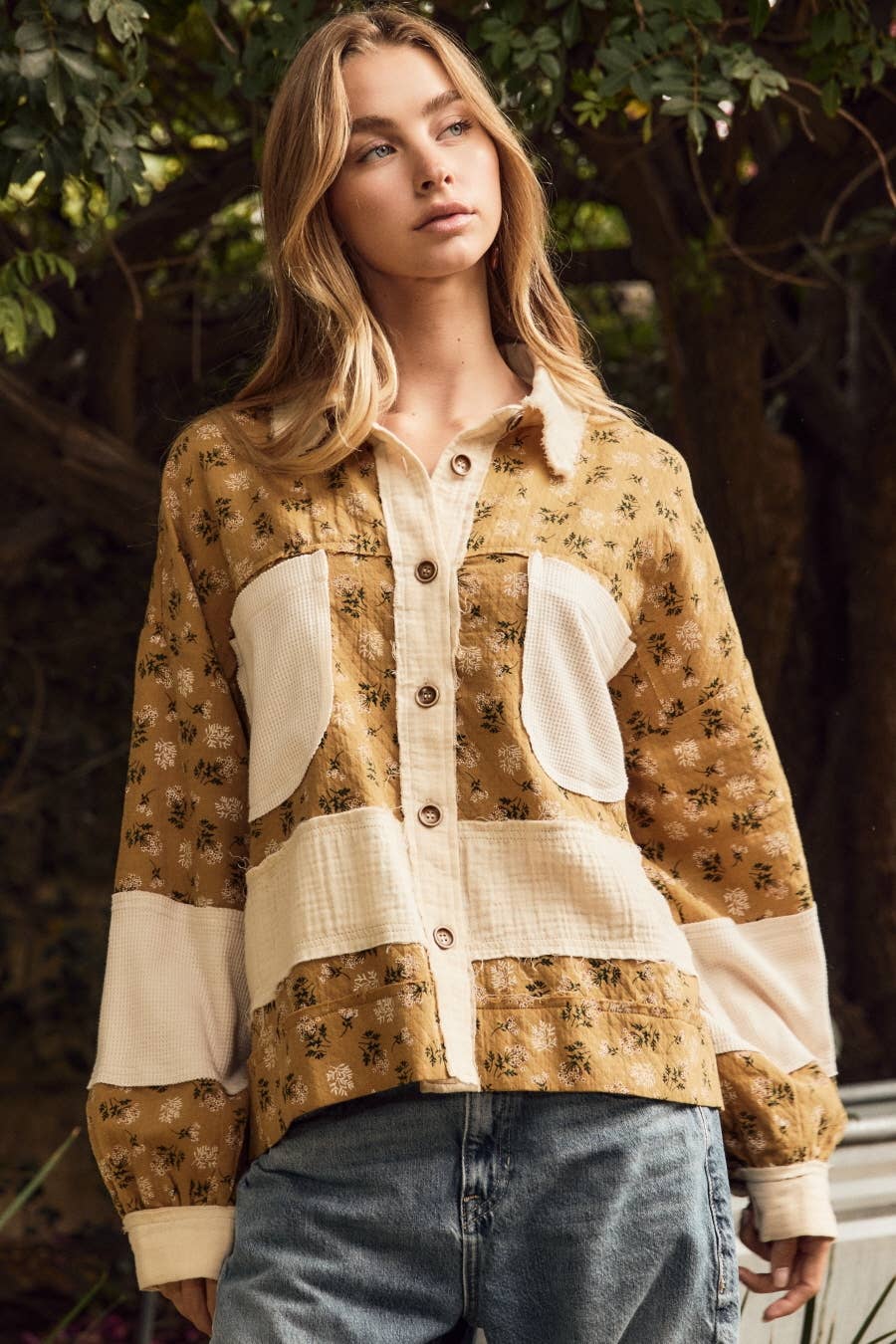 A woman wearing a patchwork button-up shirt with a tan floral print and cream-colored panels, paired with blue jeans, set against a background of green foliage.