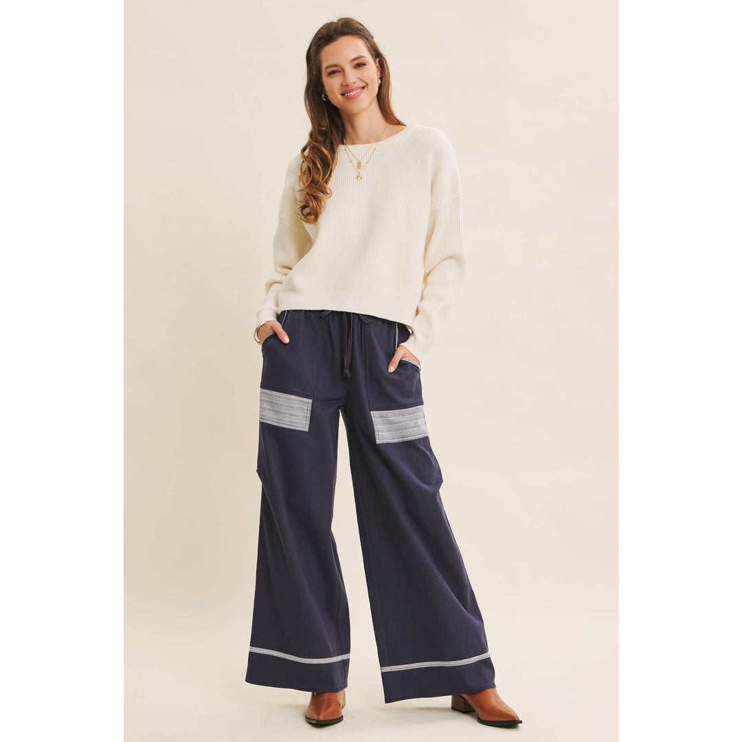 Model wearing Bow Back Sweater | Dolman Sleeve Knit with Satin Tie paired with navy pants with white lace patches and striped cuff on beige background