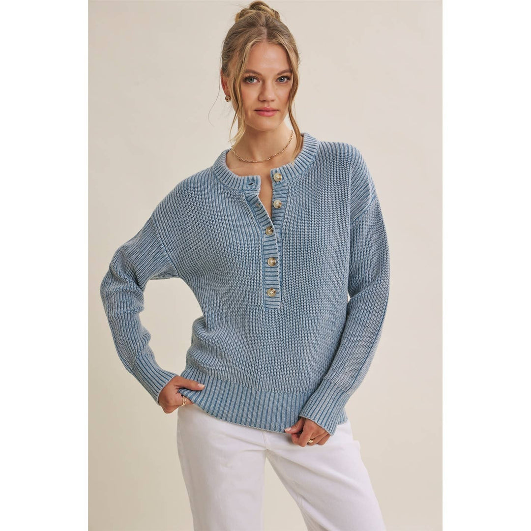 A woman modeling a light blue ribbed knit henley sweater with wooden buttons, paired with white pants, against a plain beige studio background.
