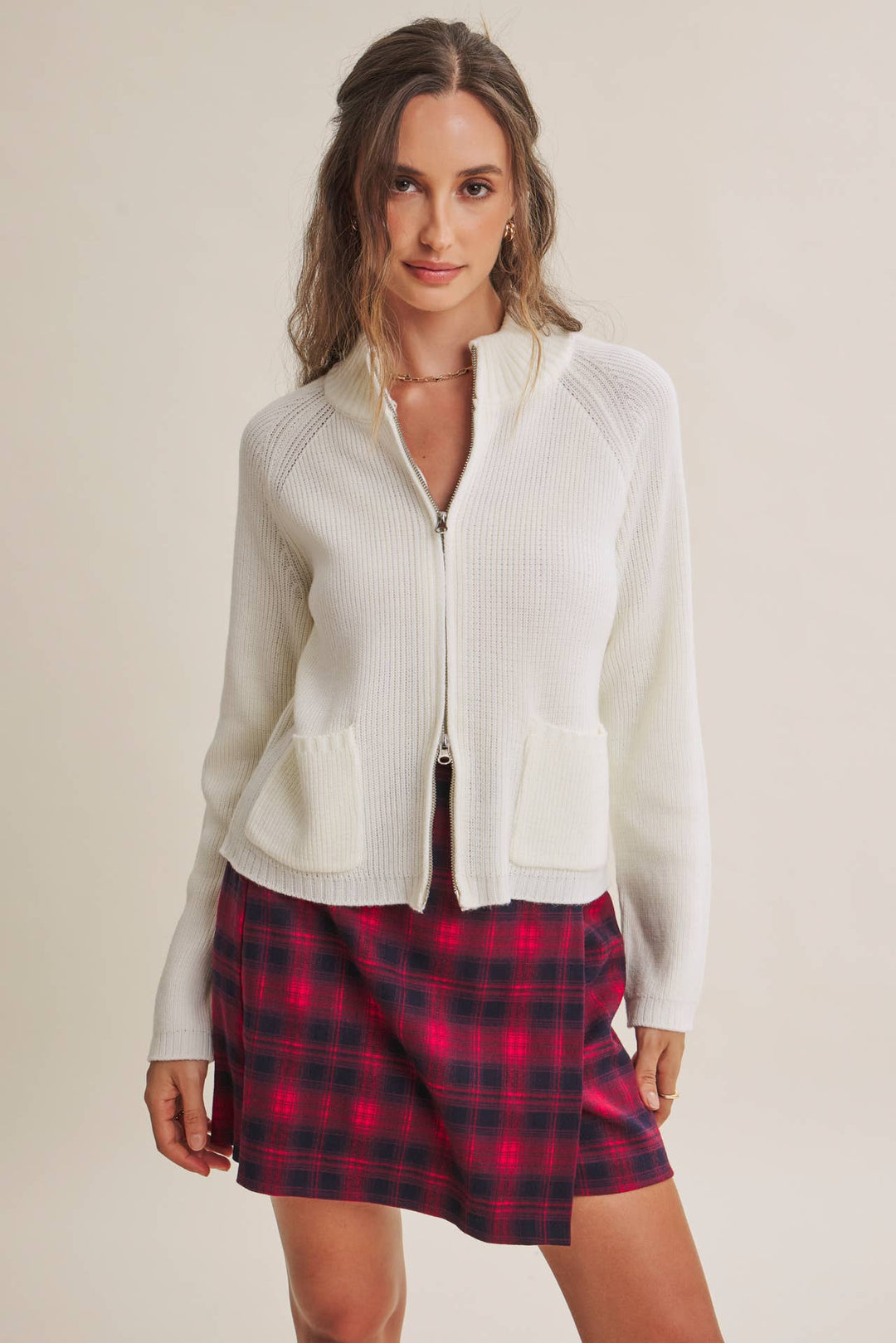 A woman modeling a white ribbed zip-up cardigan with a stand collar and front patch pockets, paired with a red and black plaid mini skirt, against a plain beige studio background.