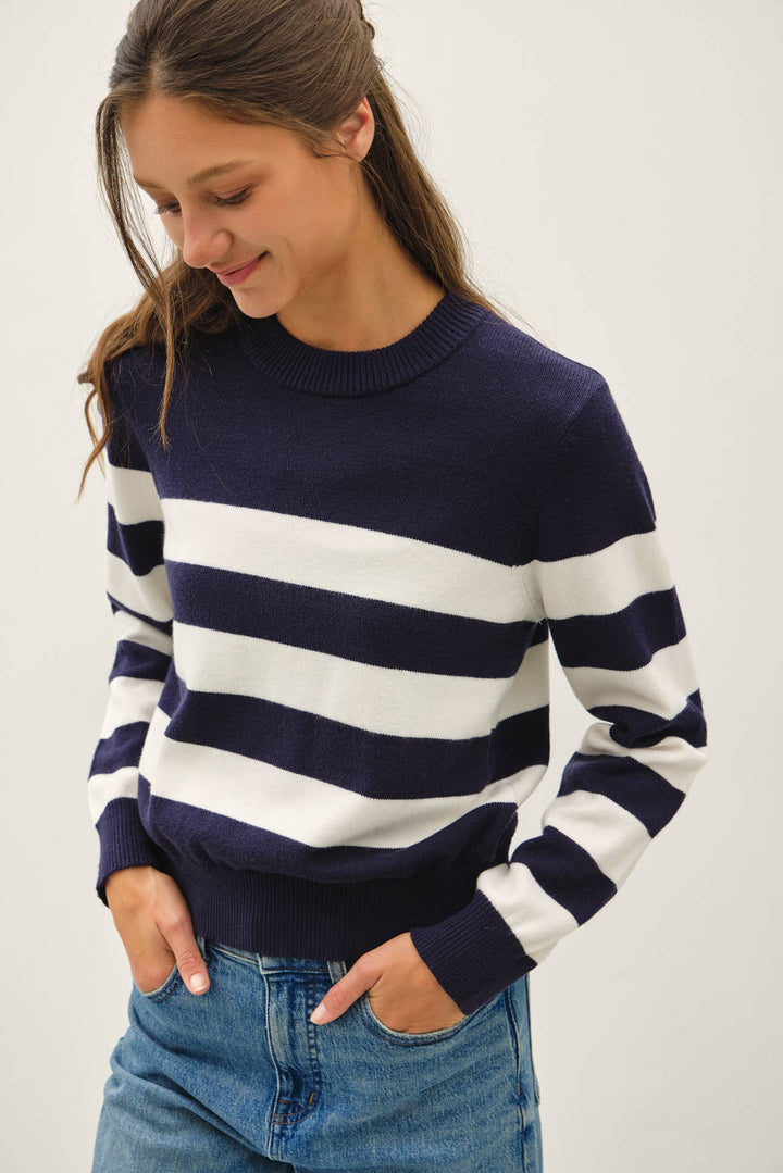 A woman wearing a navy blue and white horizontally striped knit sweater with blue jeans, standing against a plain light gray background.