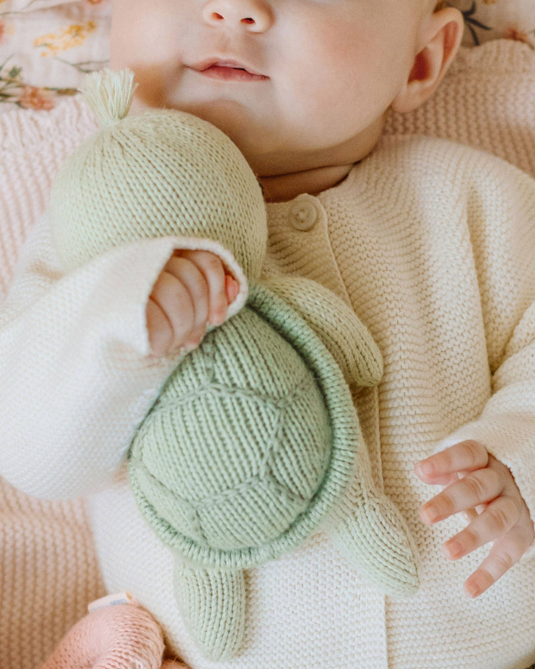 Baby holding a green knitted turtle with a soft pink blanket in the background