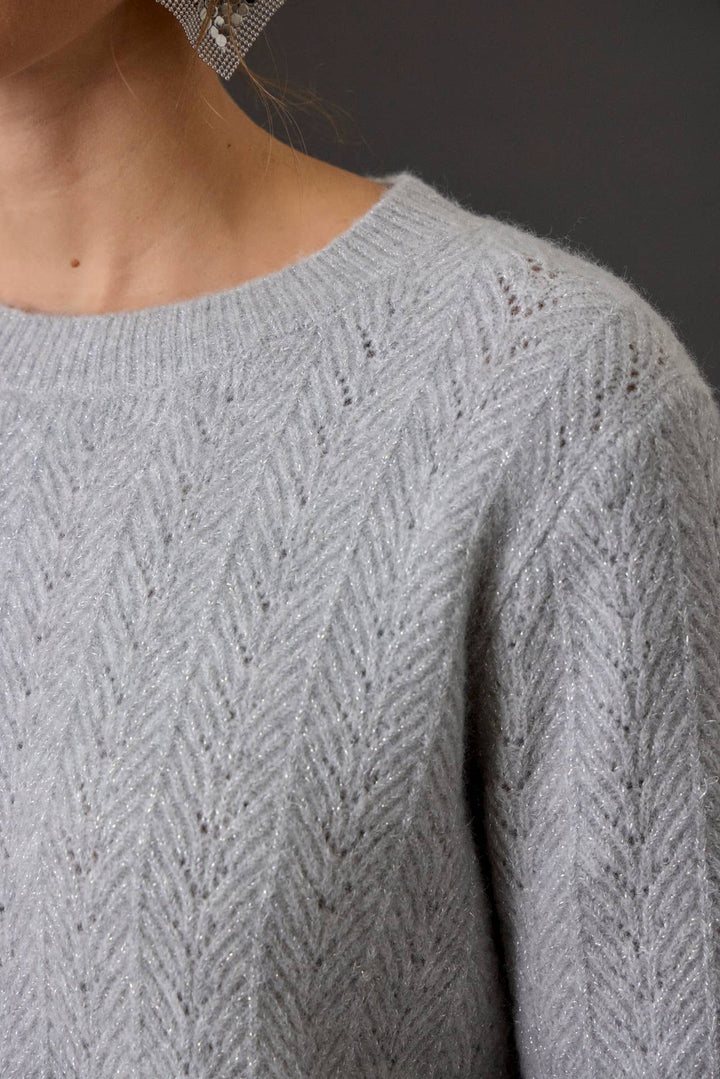 Close-up of a person wearing a light gray knit sweater with a textured herringbone pattern with shimmer threads, showing the neck, shoulder, and upper chest against a dark gray background.
