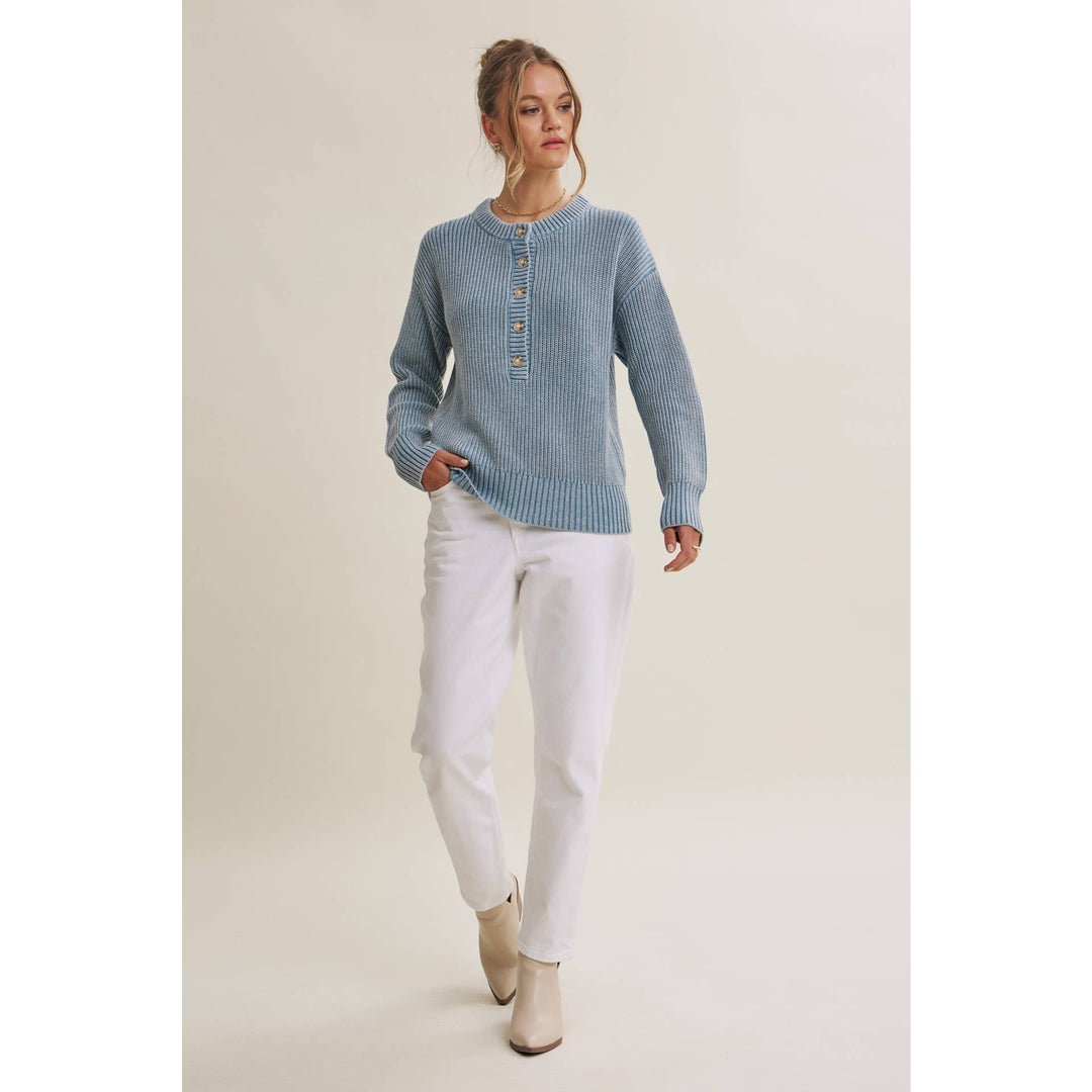 A woman modeling a light blue ribbed knit sweater with a buttoned henley neckline, paired with white pants and beige ankle boots, against a plain beige studio background.
