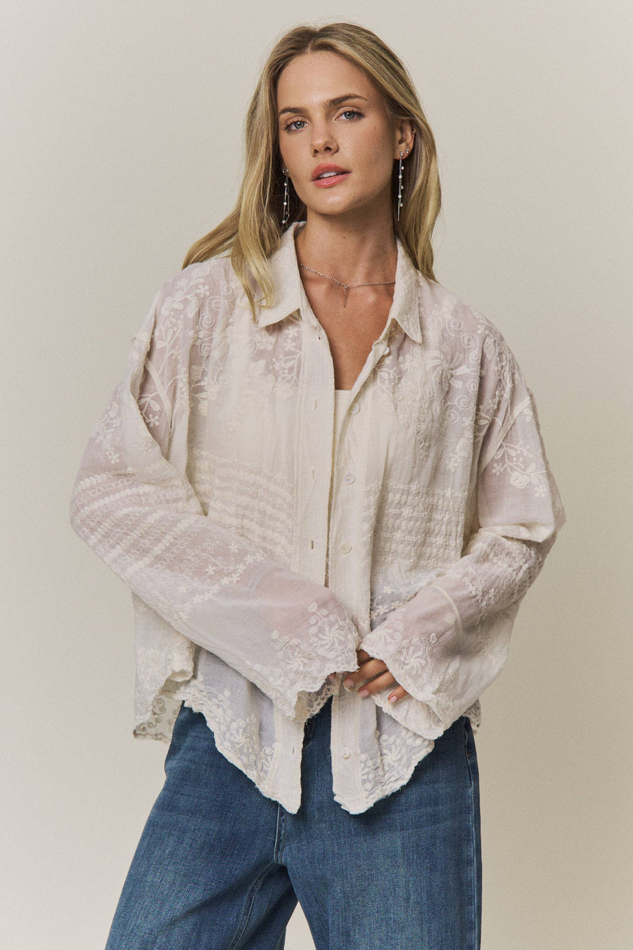 Woman wearing a light-colored lace embroidered blouse and blue jeans against a plain background