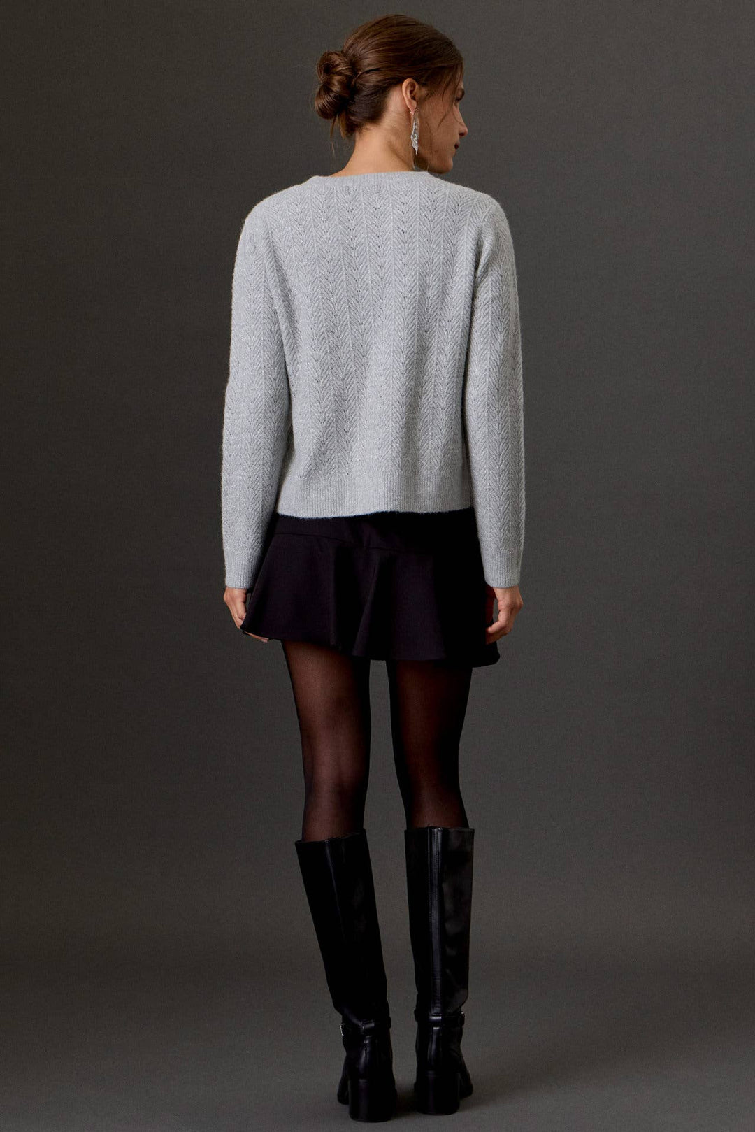 A woman shown from the back wearing a light gray knit sweater, a dark pleated mini skirt, sheer black tights, and black knee-high boots against a dark gray studio background.
