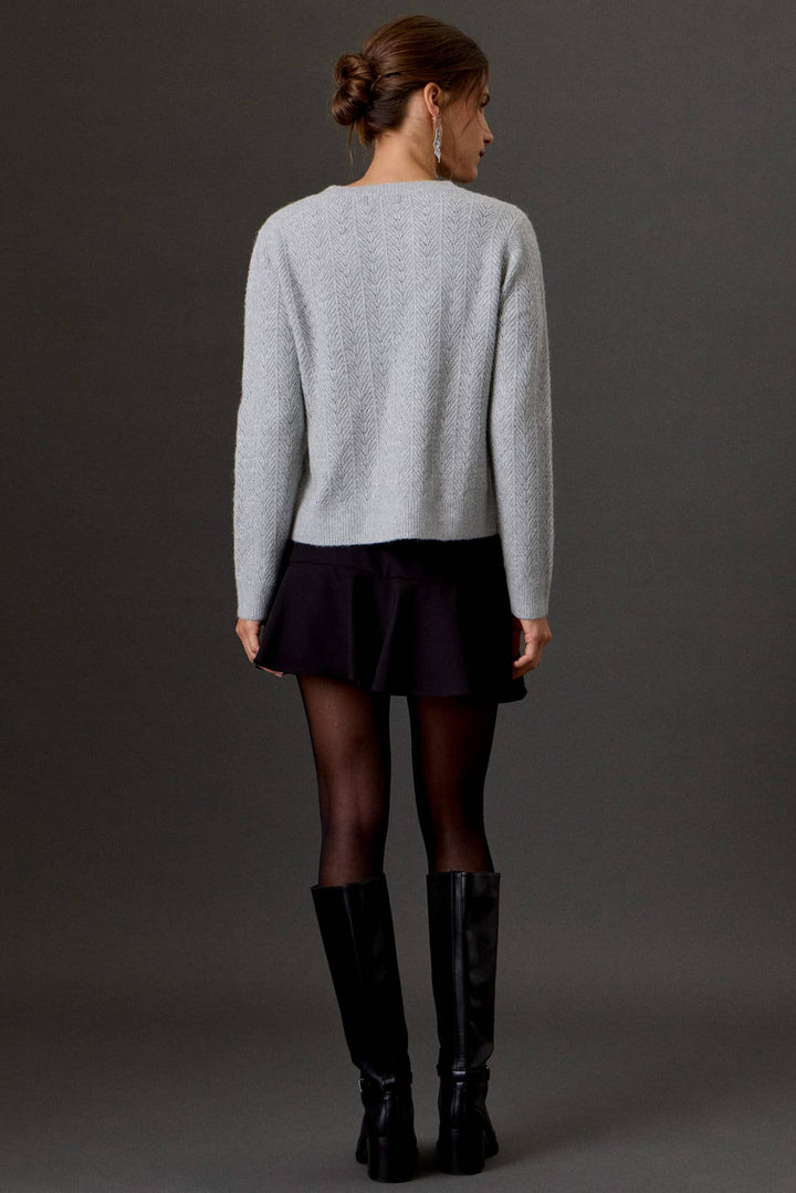 A woman shown from the back wearing a light gray knit sweater, a dark pleated mini skirt, sheer black tights, and black knee-high boots against a dark gray studio background.