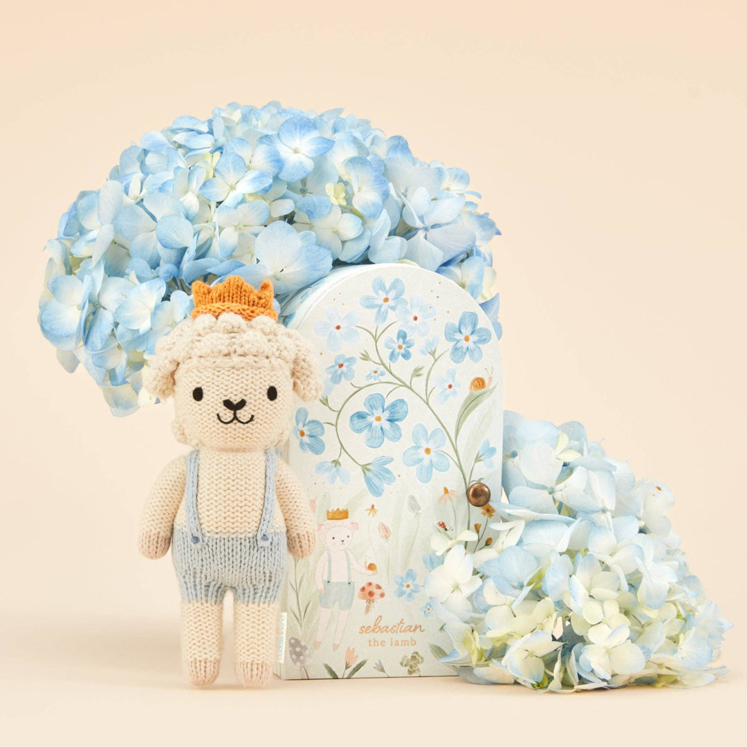Knitted lamb with a crown, surrounded by blue flowers and a decorative box on a beige background