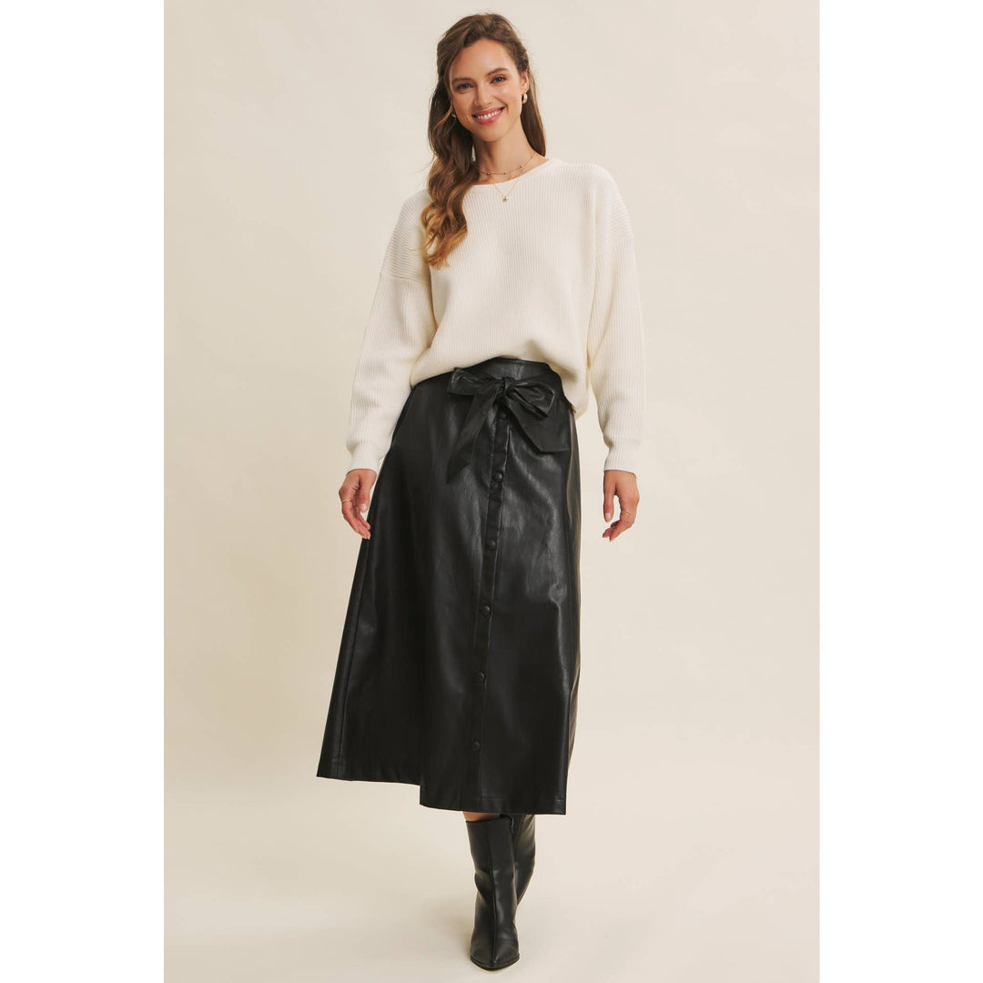 Woman wearing a Bow Back Sweater | Dolman Sleeve Knit with Satin Tie paired with a faux black leather skirt on a beige background