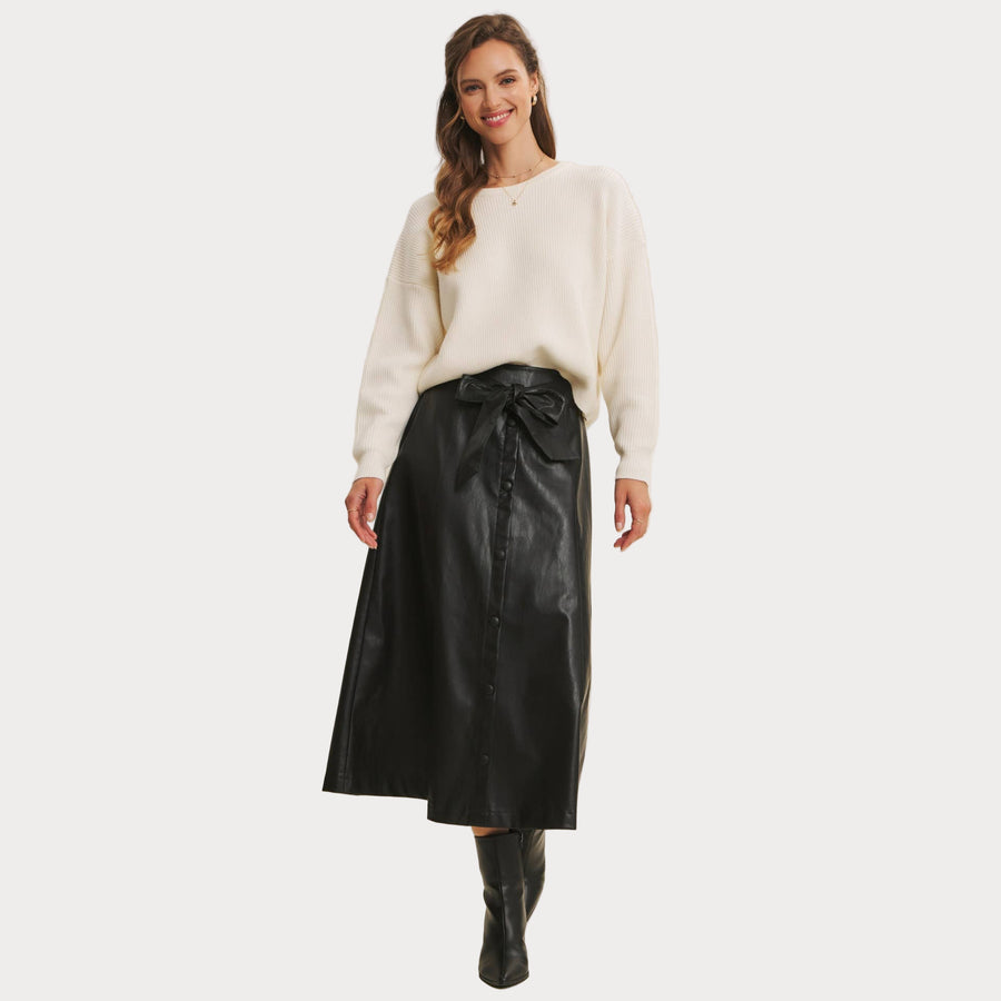 Woman wearing a Bow Back Sweater | Dolman Sleeve Knit with Satin Tie paired with a faux black leather skirt on a beige background