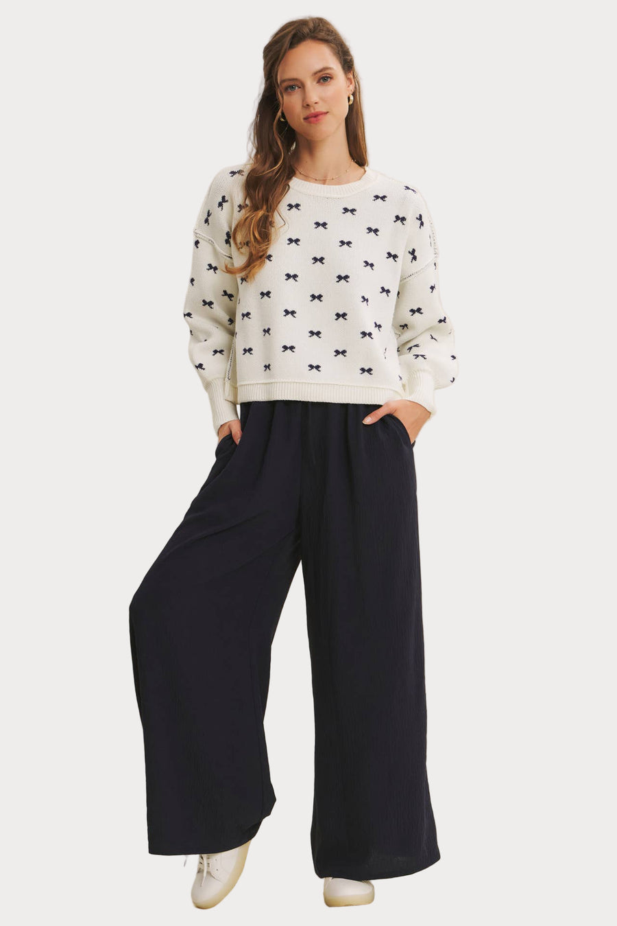 Model wearing an ivory jacquard knit sweater with a repeating navy blue bow pattern and voluminous balloon sleeves. Paired with wide-leg navy pants and white sneakers, styled for a casual, chic look.
