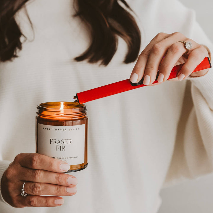 Person lighting a Fraser Fir candle with a red lighter, wearing a white sweater.