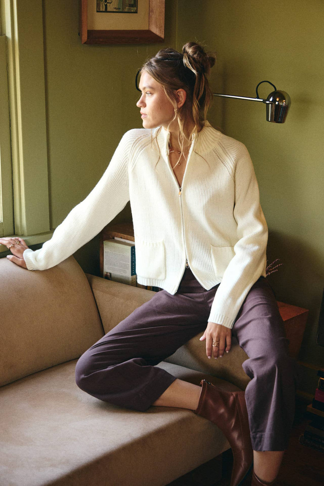 Woman sitting on a couch wearing a white jacket and purple pants in a room with green walls.