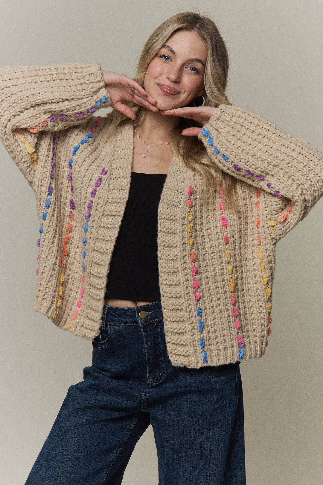 A woman wearing a chunky knit cardigan in beige with colorful yarn-like vertical stripes in pink, blue, yellow, and purple, styled over a black top and dark blue jeans against a plain beige studio background.