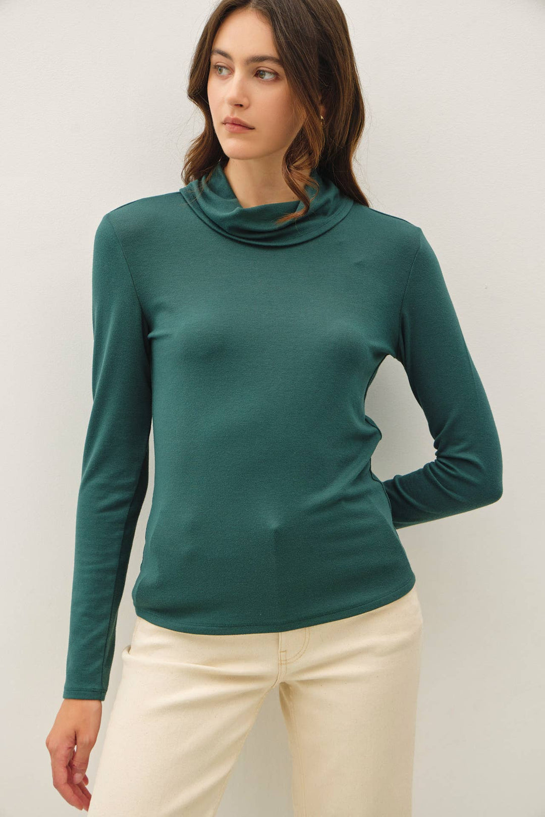 A woman wearing a dark green long-sleeve turtle neck top and cream pants, standing against a light gray studio background.