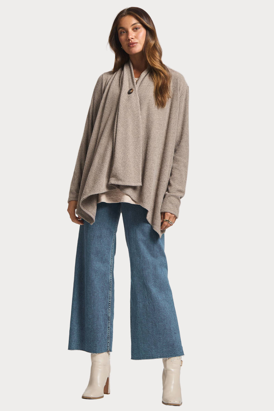 Woman wearing a beige wrap cardigan, light gray top, blue jeans, and white boots on a white background