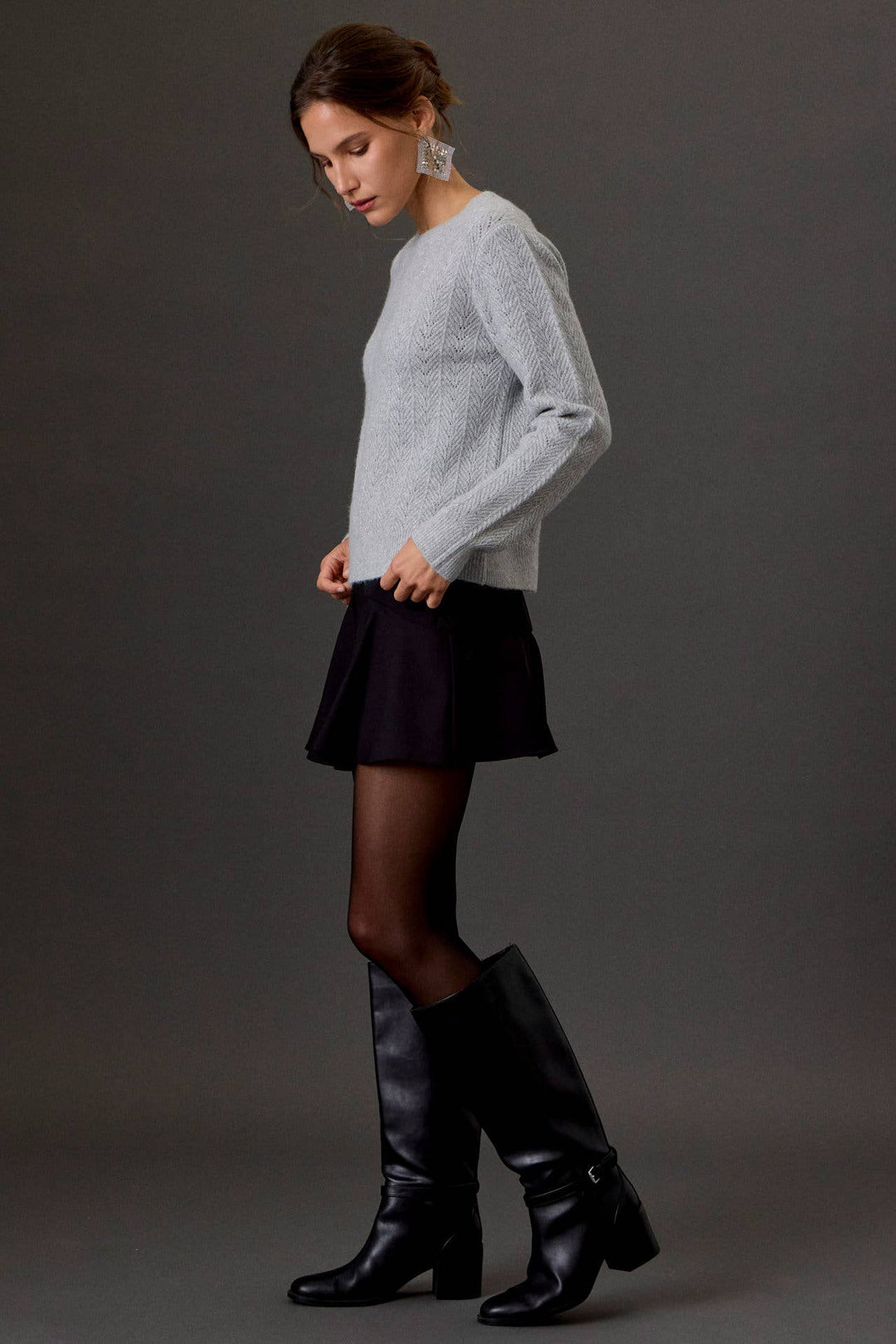 A woman models a light gray knit sweater, a black pleated mini skirt, sheer black tights, and black leather knee-high boots against a dark gray studio background.