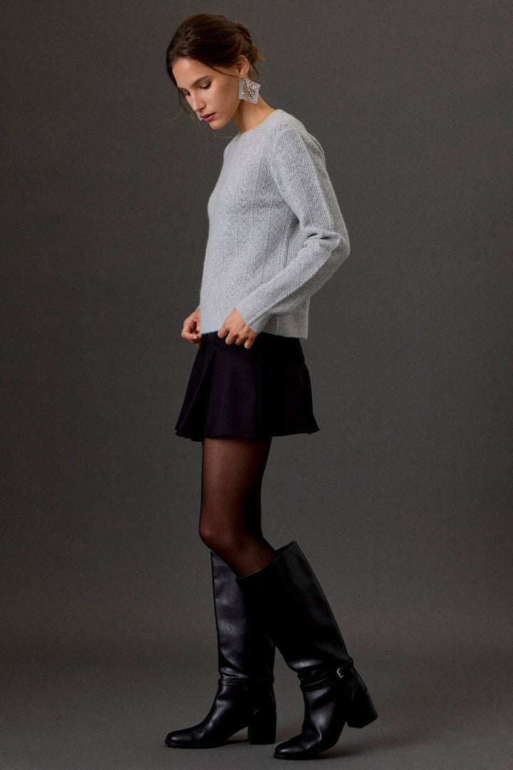 A woman models a light gray knit sweater, a black pleated mini skirt, sheer black tights, and black leather knee-high boots against a dark gray studio background.