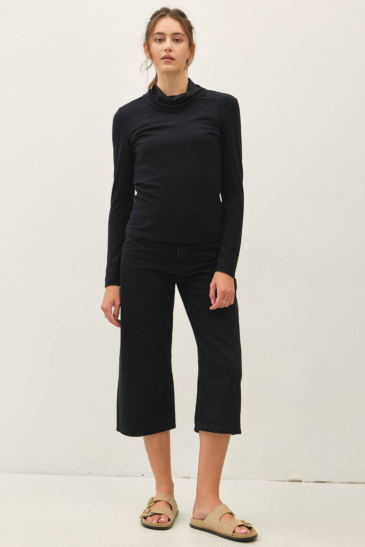 A woman modeling a black long-sleeve turtle neck top and matching black cropped pants, paired with beige slide sandals, against a plain off-white studio background.