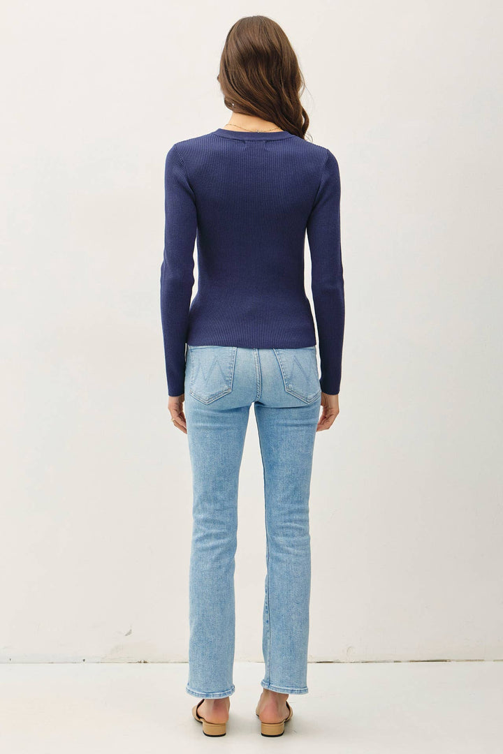 A woman viewed from behind wearing a navy blue long-sleeve top and light-wash blue jeans, standing against a plain off-white studio background.
