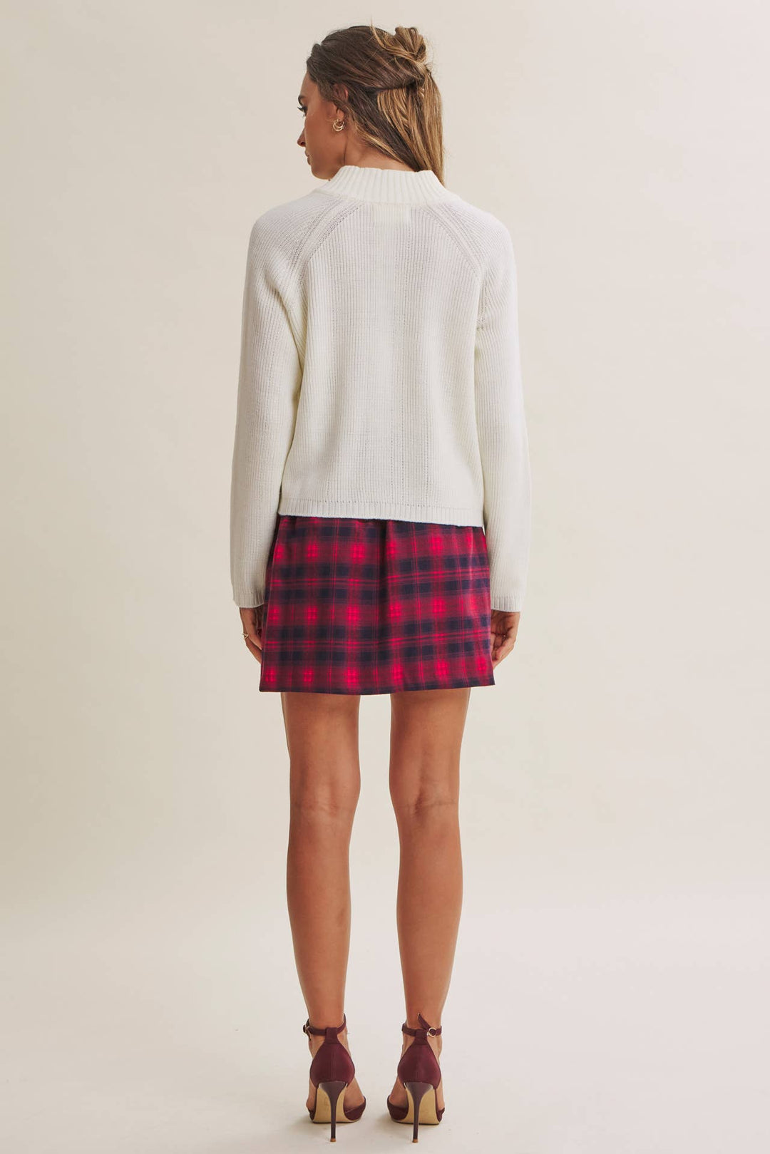 A woman shown from the back wearing a cream mock-neck knit sweater, a red and black plaid mini skirt, and burgundy high-heeled sandals, set against a plain light beige studio background.