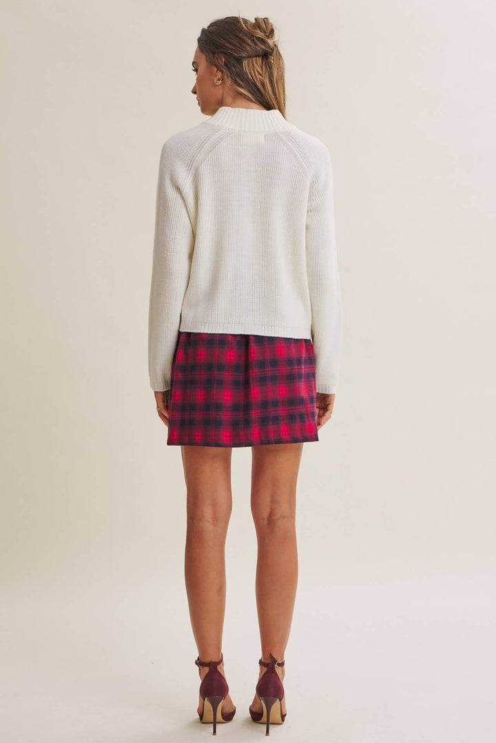 A woman shown from the back wearing a cream mock-neck knit sweater, a red and black plaid mini skirt, and burgundy high-heeled sandals, set against a plain light beige studio background.