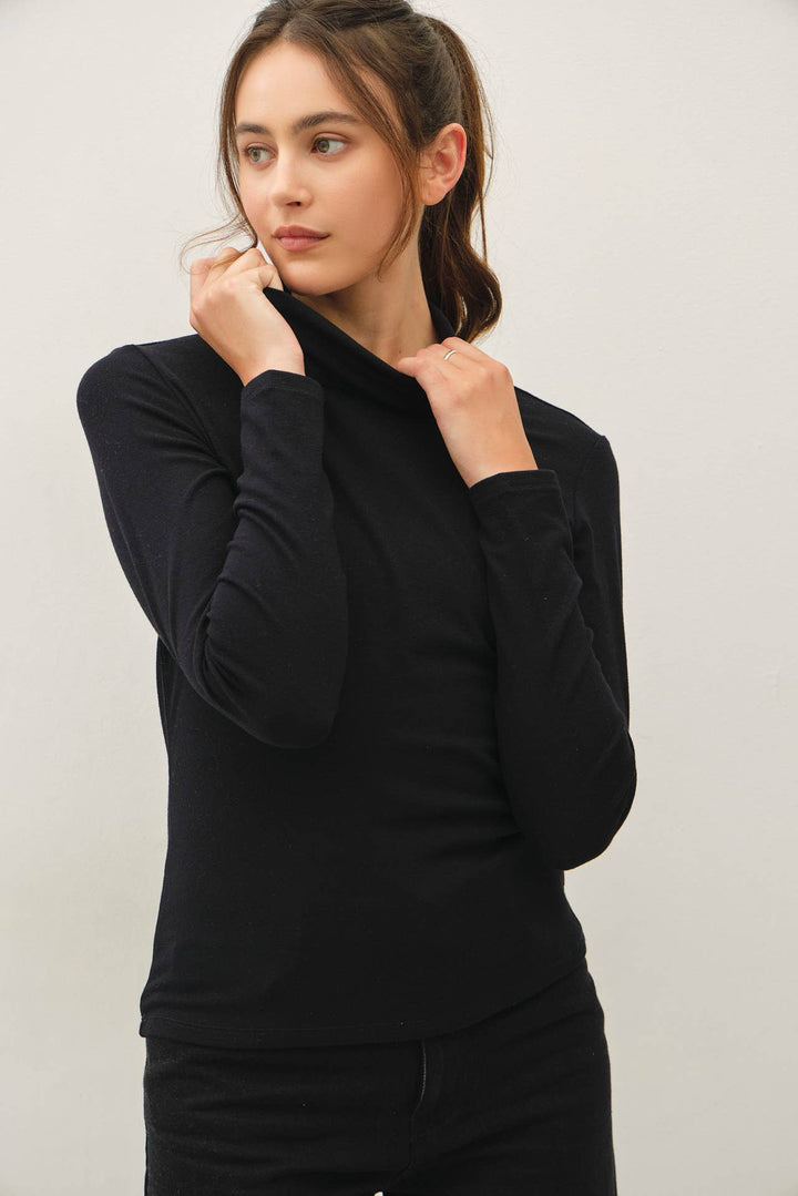 A woman wearing a black long-sleeve turtle neck top, posing with her hands near the collar against a plain light gray background.
